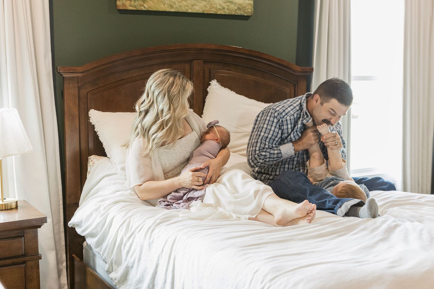 Family with newborn baby sitting on a white bed in a cozy bedroom with green walls and wooden headboard.