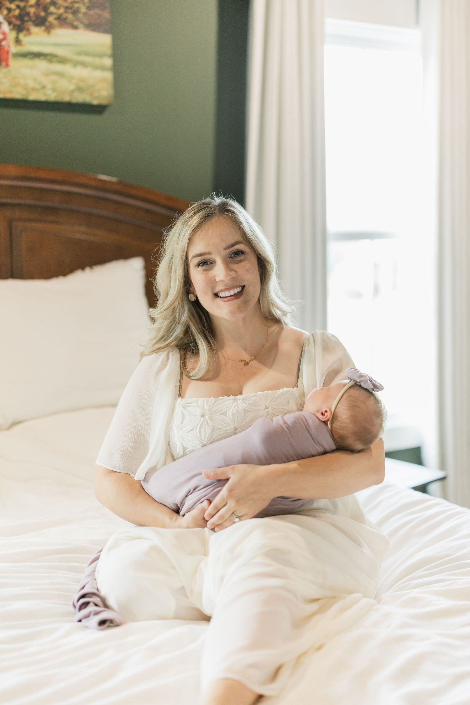 Smiling mother sitting on white bed holding swaddled newborn baby in cozy bedroom setting.