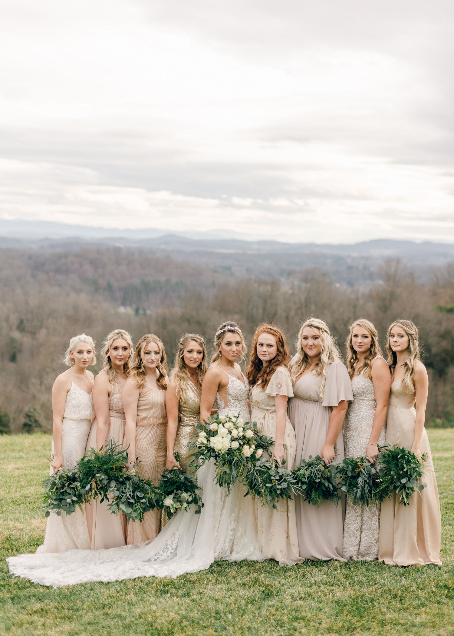 Bride and bridesmaids in champagne gowns holding greenery bouquets outdoors with rolling hills backdrop.