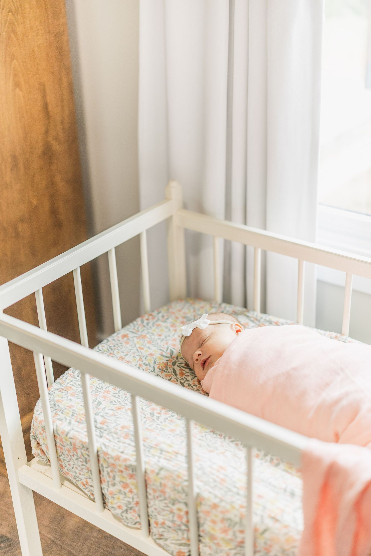 Newborn baby sleeping peacefully in a white crib with floral sheets, wrapped in a pink swaddle blanket.