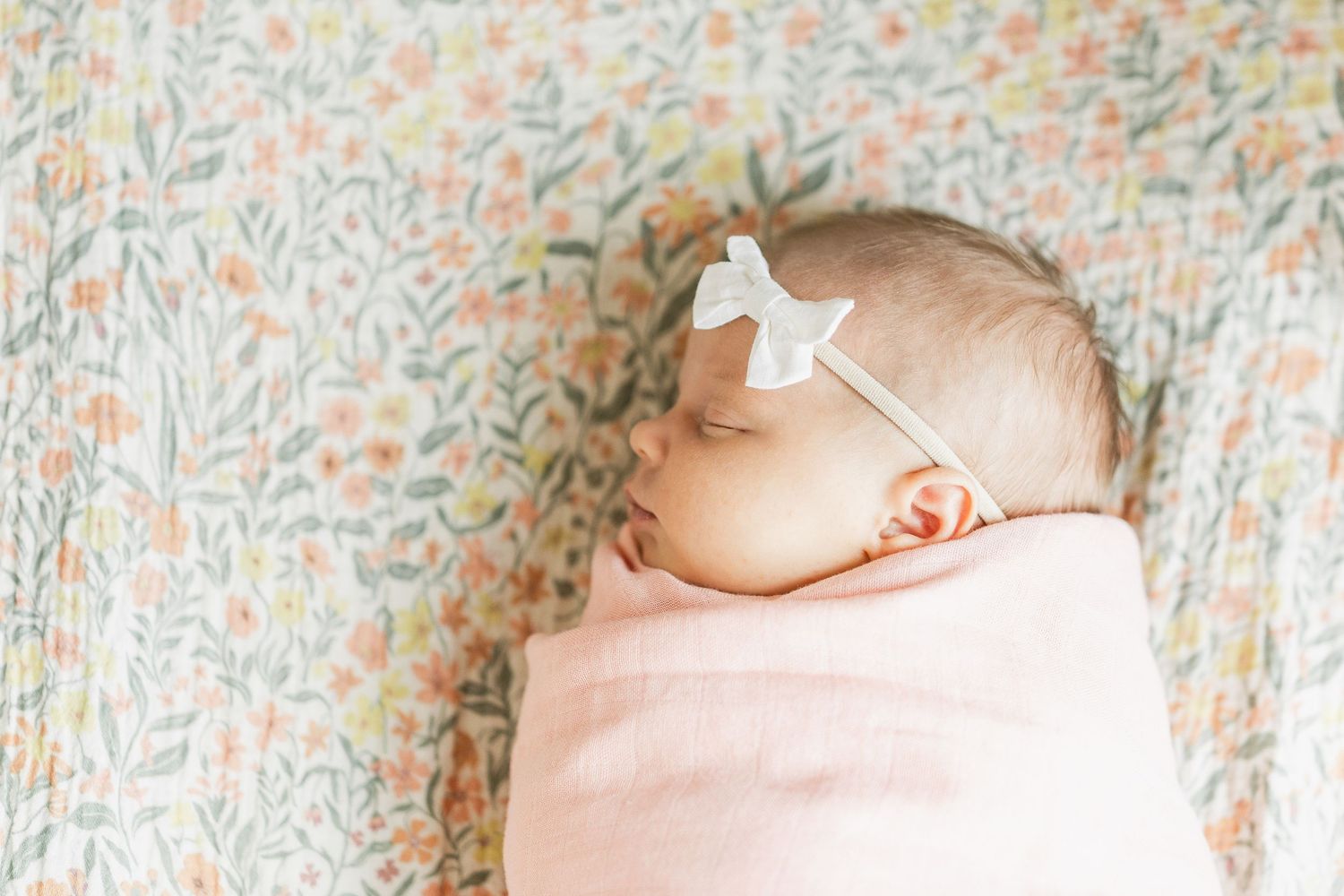 Sleeping newborn baby swaddled in pink blanket with white bow headband on floral background.