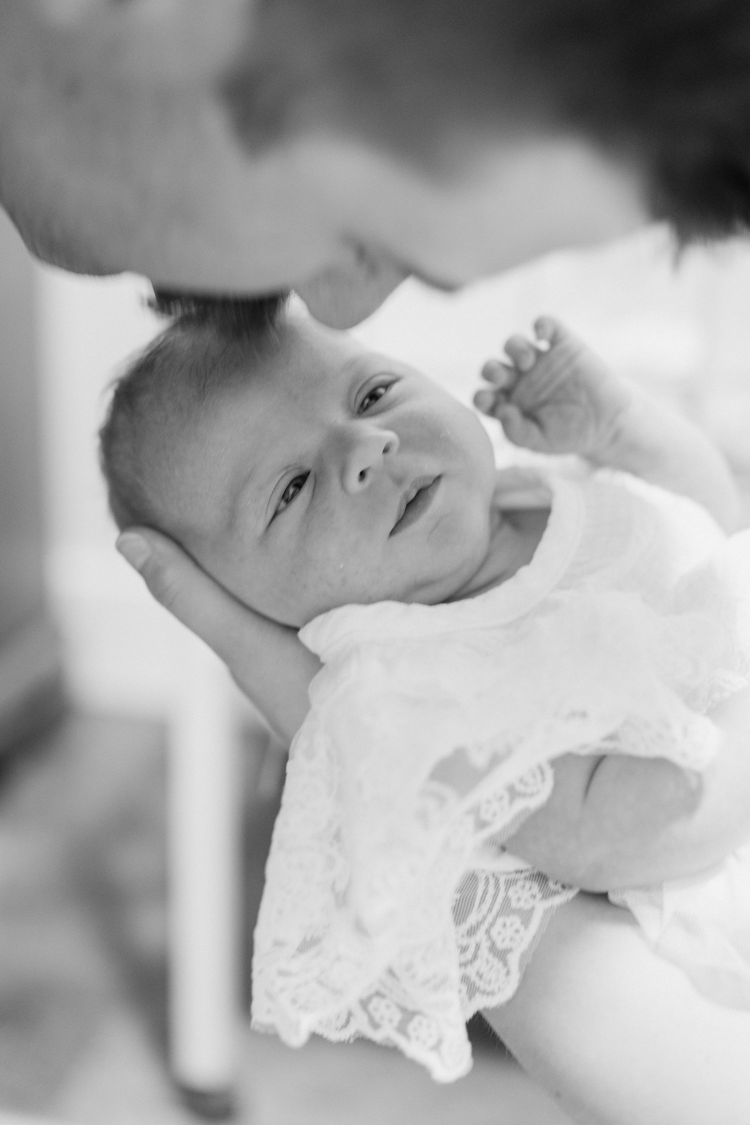 Black and white photo of a newborn baby in lace outfit being held and kissed by mother.
