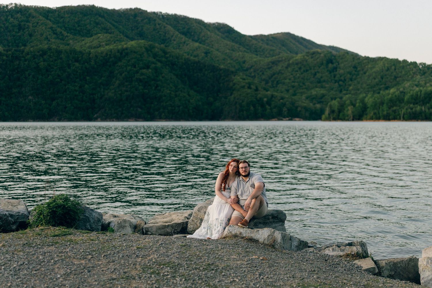 Couple sitting together on rocks by a lake, surrounded by lush green mountains in soft evening light.