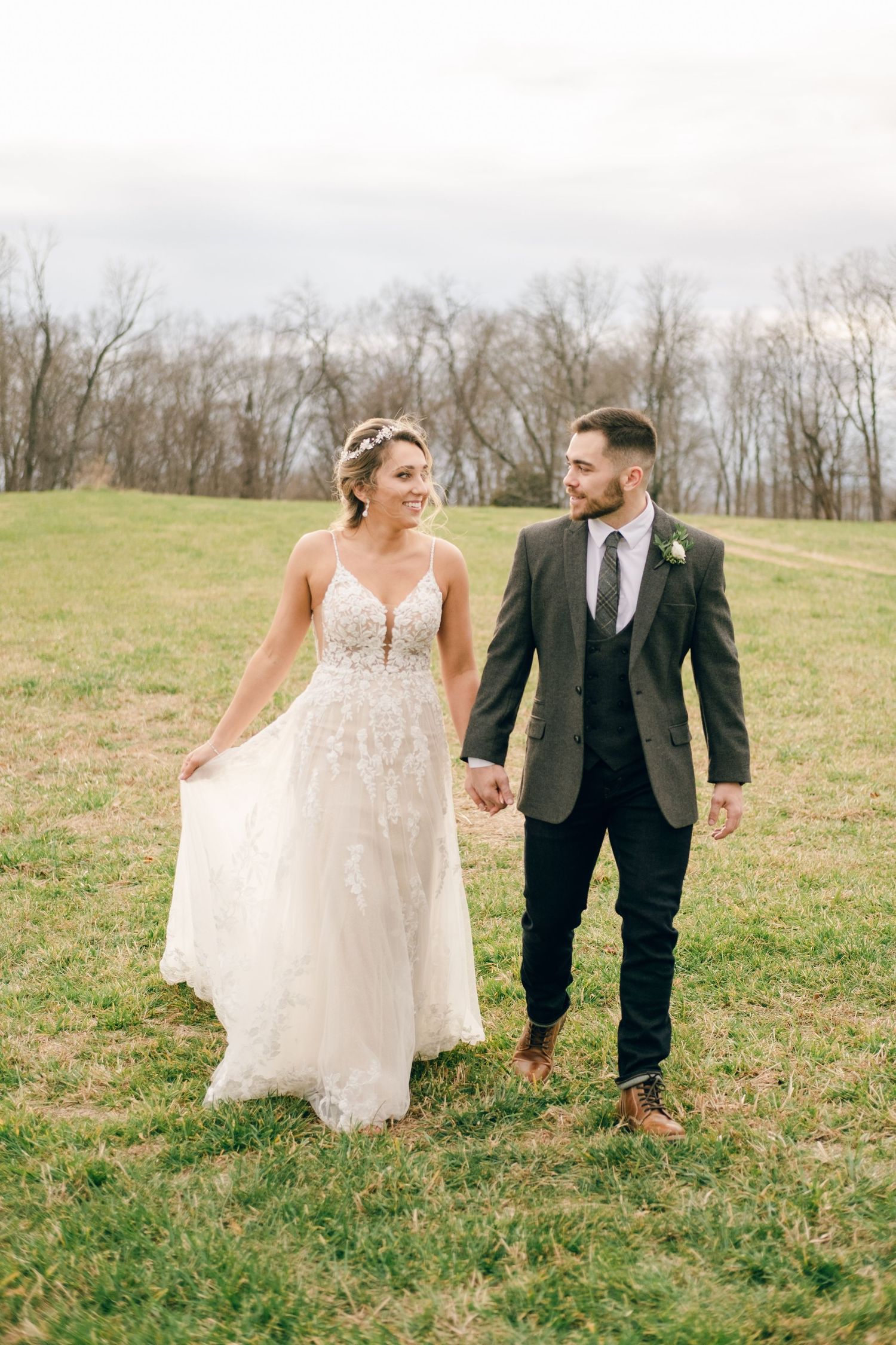 A bride and groom walk hand-in-hand through a green field, smiling at each other on their wedding day.