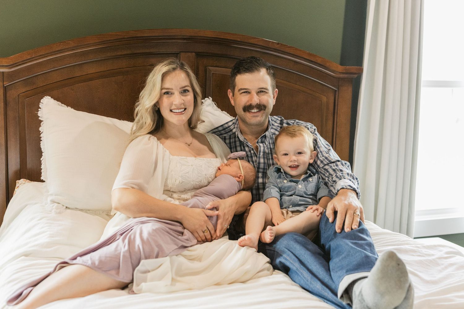 A happy family of four poses on a bed, with parents smiling and holding their newborn and toddler in a cozy bedroom.