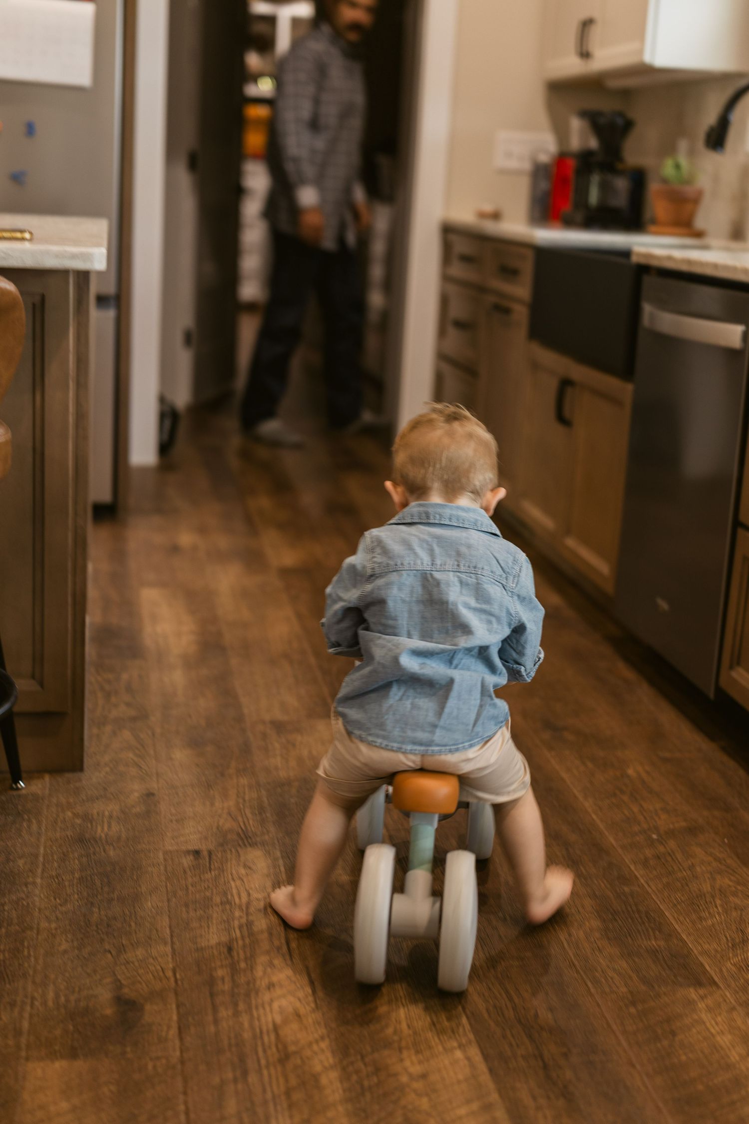 Toddler in denim shirt rides toy tricycle on hardwood floor toward adult in modern kitchen.