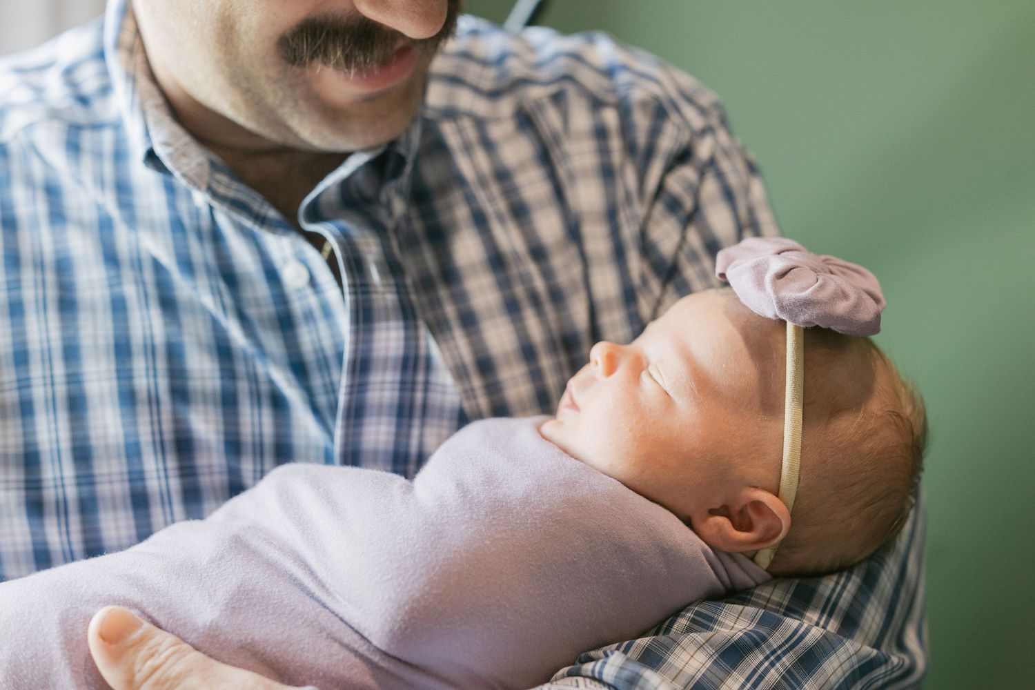 A father with a mustache tenderly holds his sleeping newborn baby girl wrapped in pink with a floral headband.