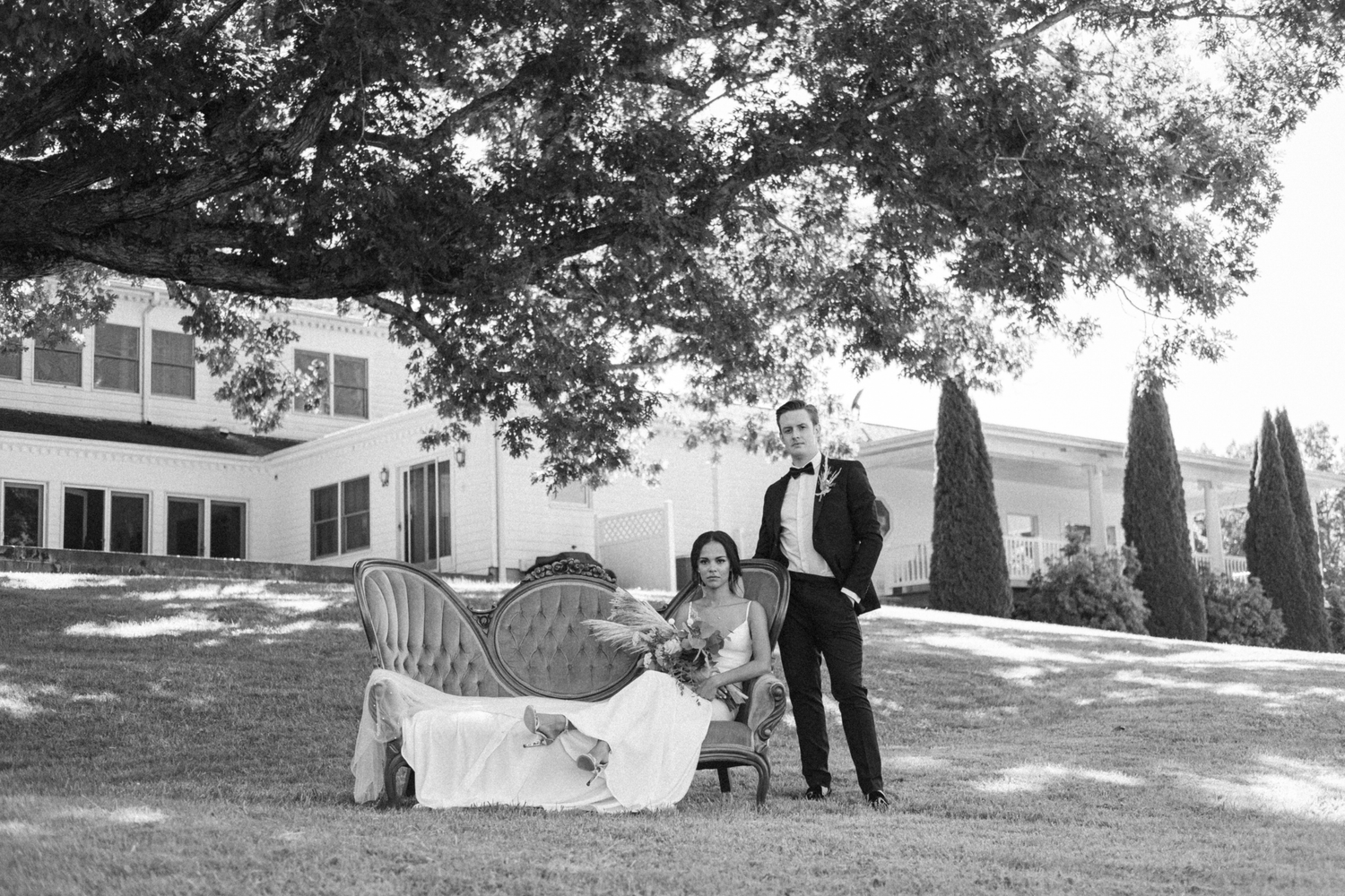 Black and white photo of bride and groom posing outdoors on lawn with vintage sofa under large tree near elegant estate.