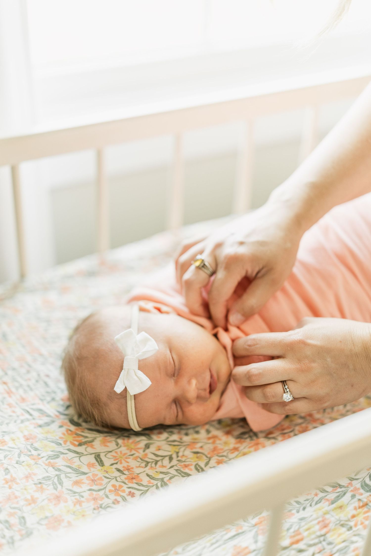Newborn baby girl in peach swaddle with white bow headband sleeping peacefully in a floral crib.