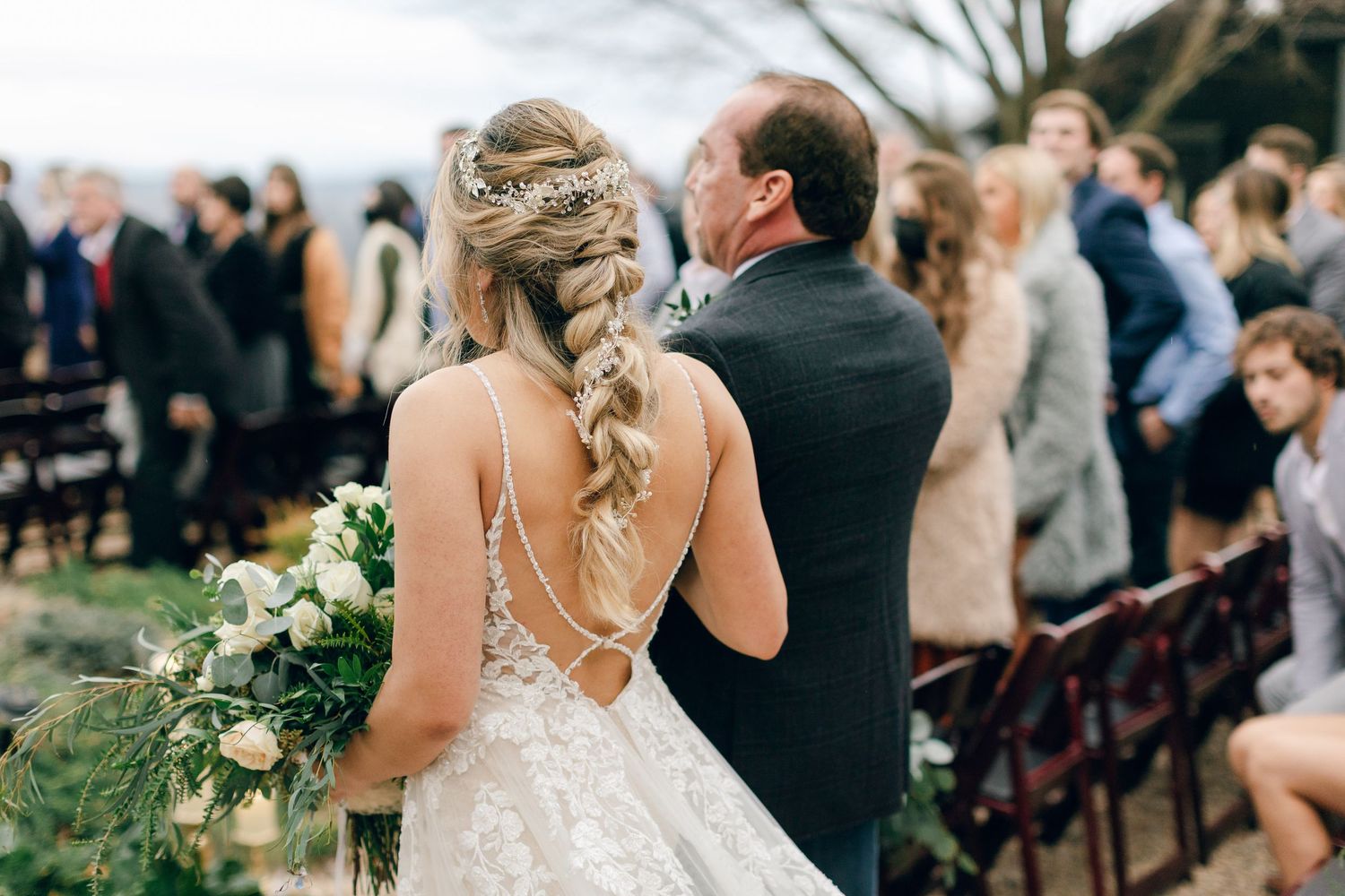 Bride with braided hair and floral crown walks down outdoor aisle holding bouquet, escorted by man in dark suit.