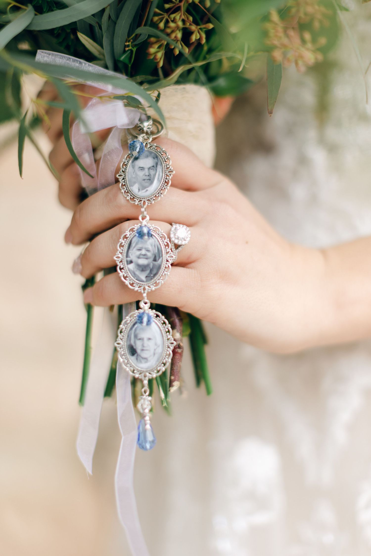 Bride holds bouquet with silver oval photo memorial charms and blue crystal pendants honoring loved ones on wedding day.