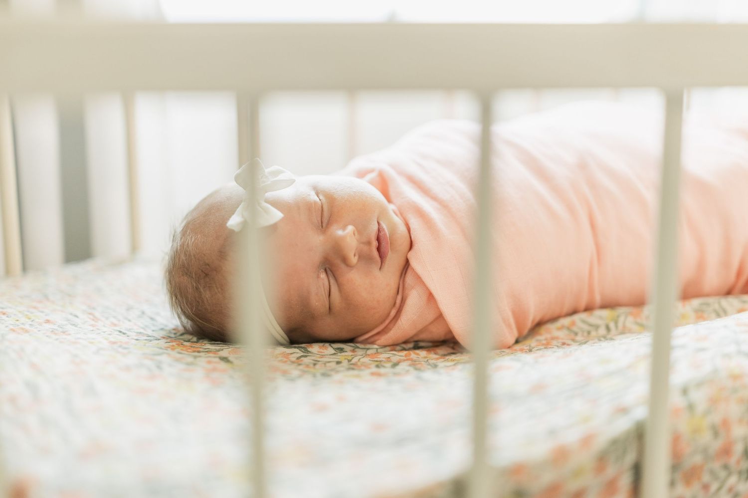 Newborn baby swaddled in pink sleeping peacefully in a white crib with floral mattress.