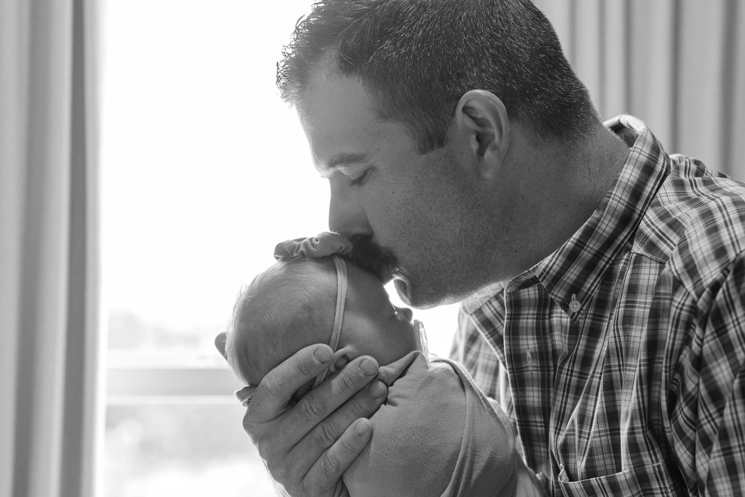 Black and white photo of a father tenderly kissing his newborn baby while cradling it gently in his hands.