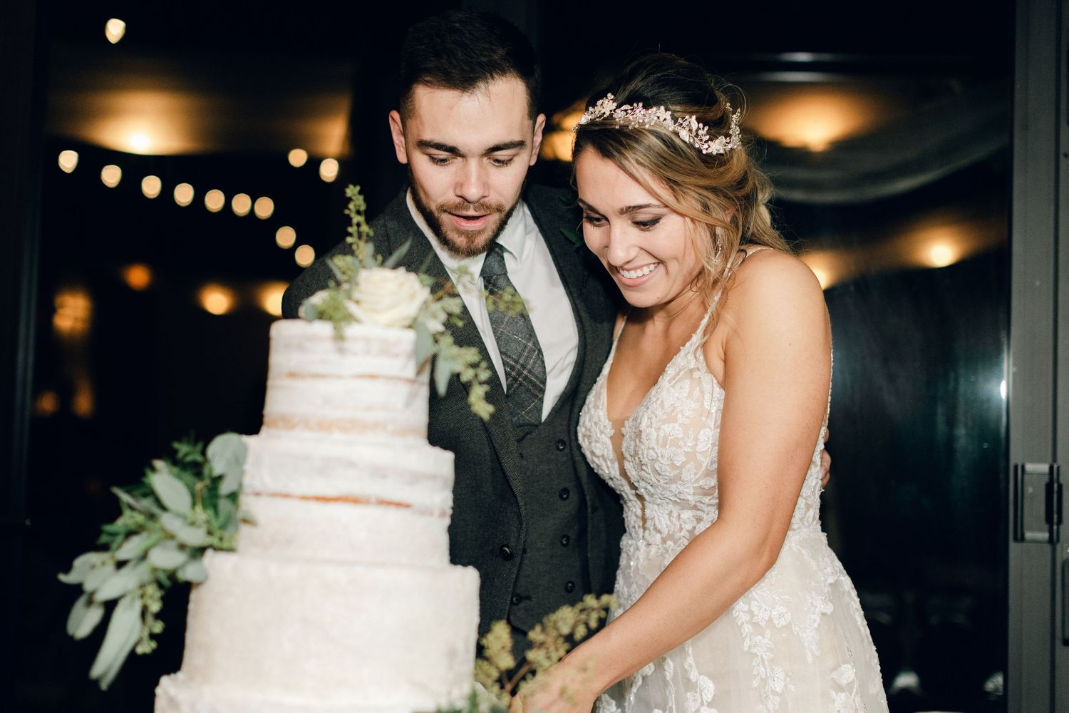 Bride and groom cutting a white four-tier wedding cake adorned with greenery at their elegant evening reception.