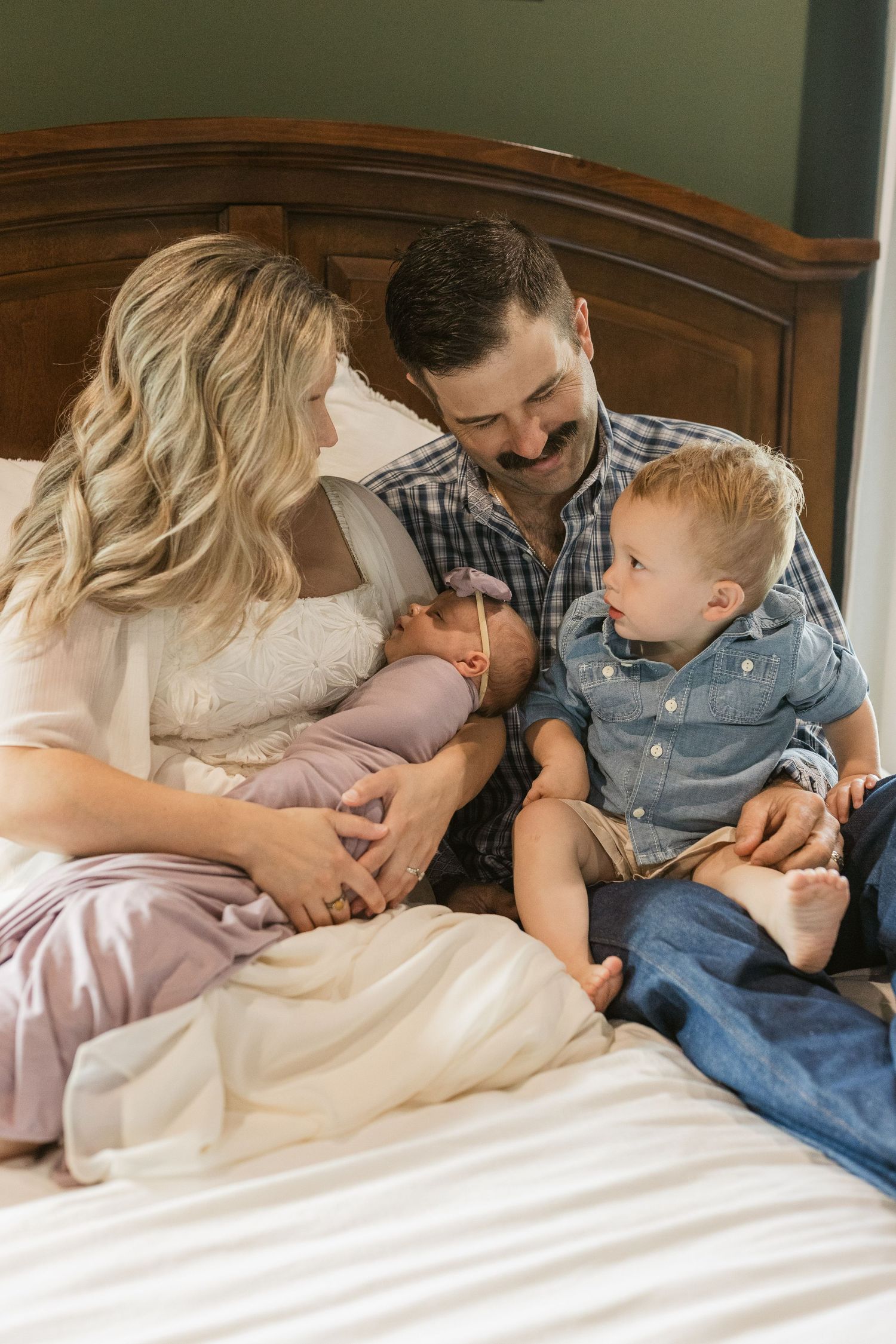 Happy family sitting on bed, mother holding newborn baby while toddler son looks on with father smiling.