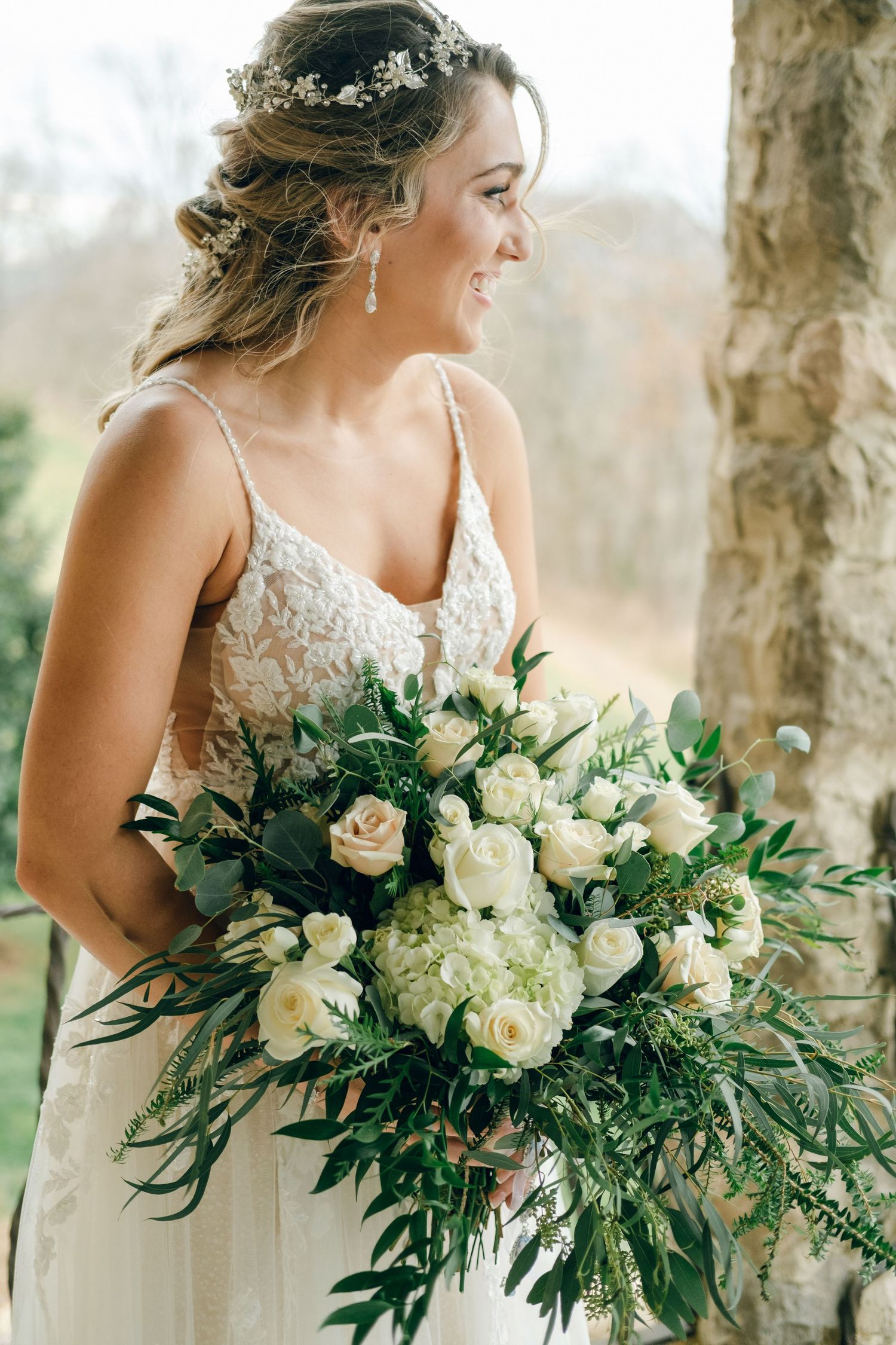 Smiling bride in lace gown holds large white rose and greenery bouquet, wearing floral hair crown outdoors.