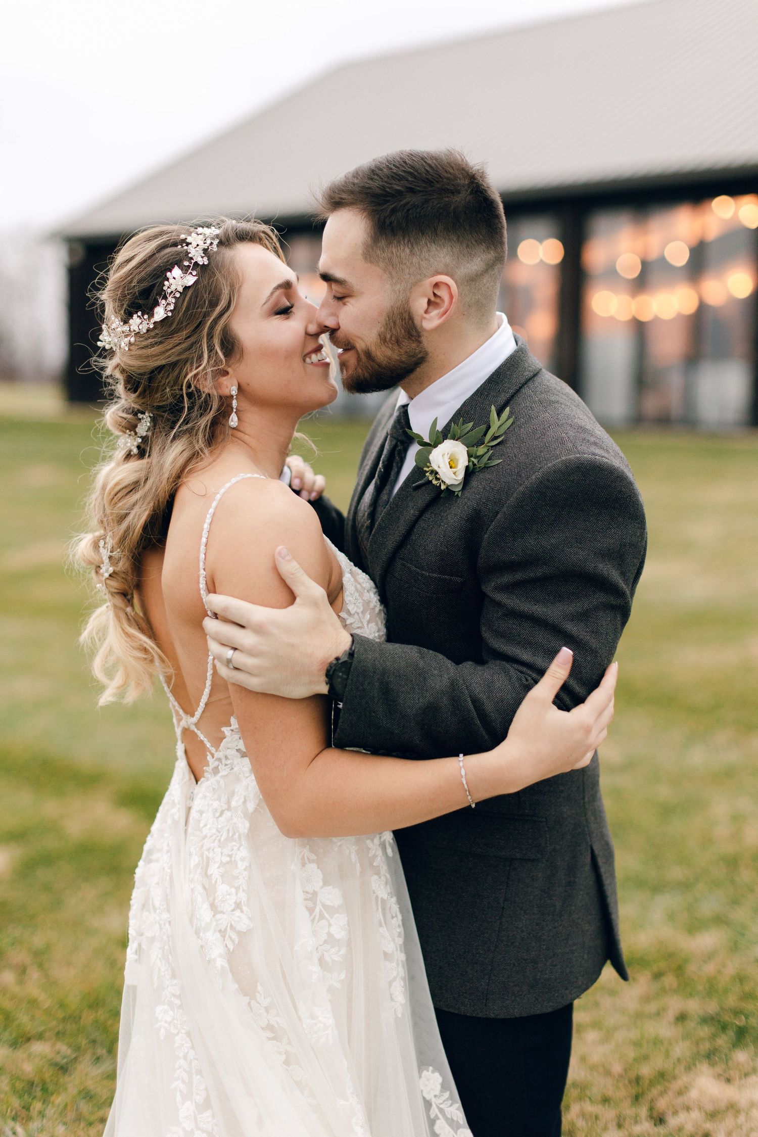 A bride and groom share a romantic almost-kiss outdoors near a barn with string lights, she in lace gown, he in gray suit.