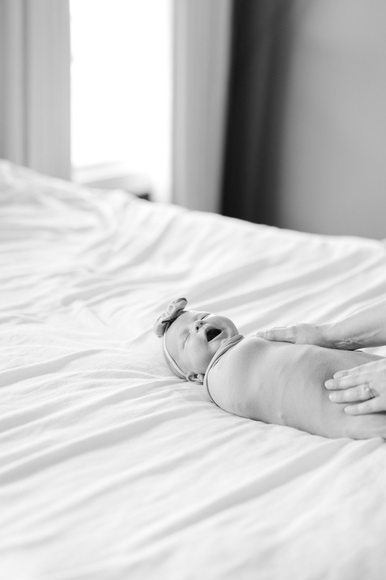Black and white photo of a newborn baby yawning while lying on white bed sheets, with adult hands gently holding them.