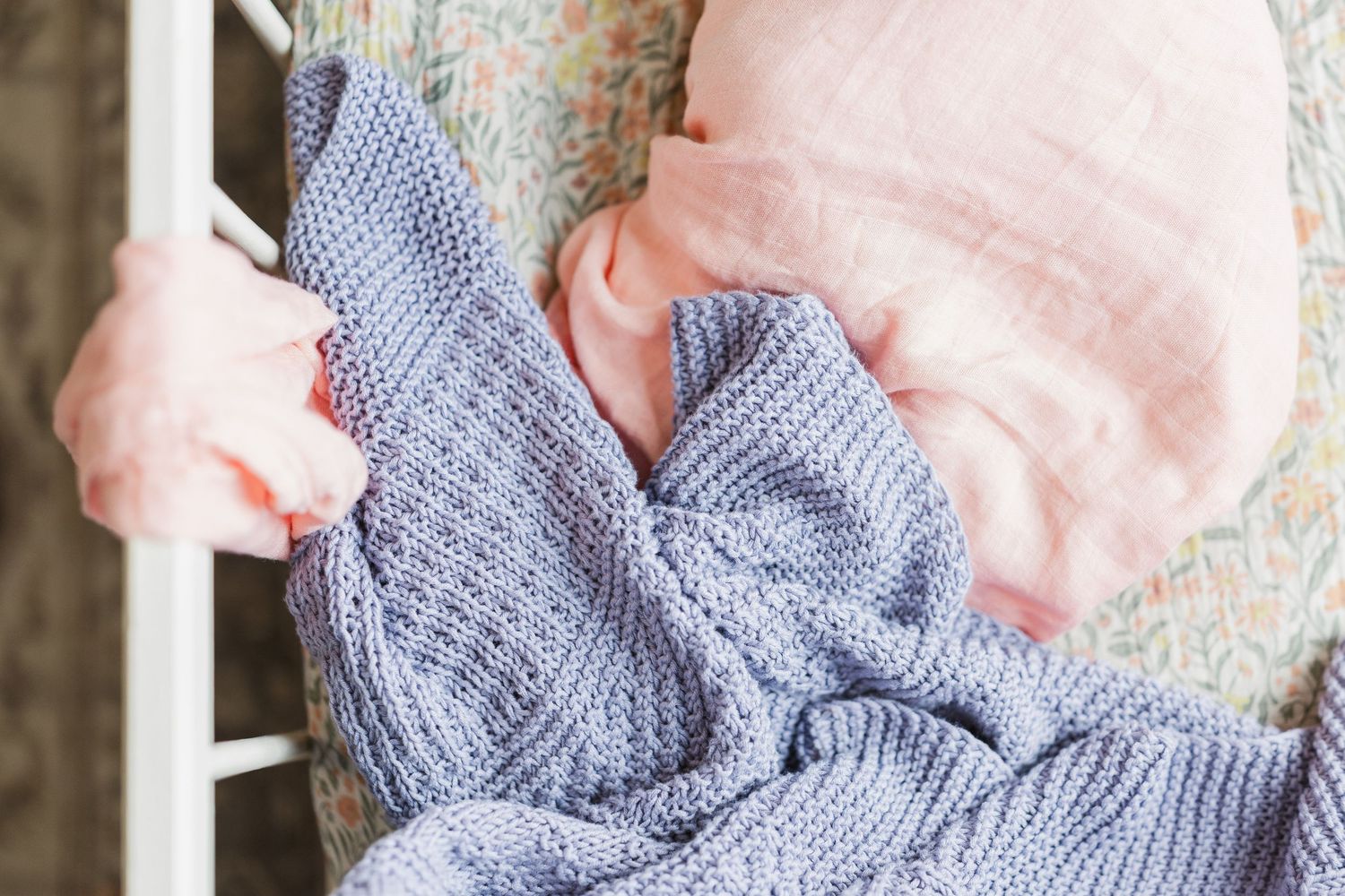 Baby sleeping in a crib wrapped in a soft blue knitted blanket with a pink pillow nearby.