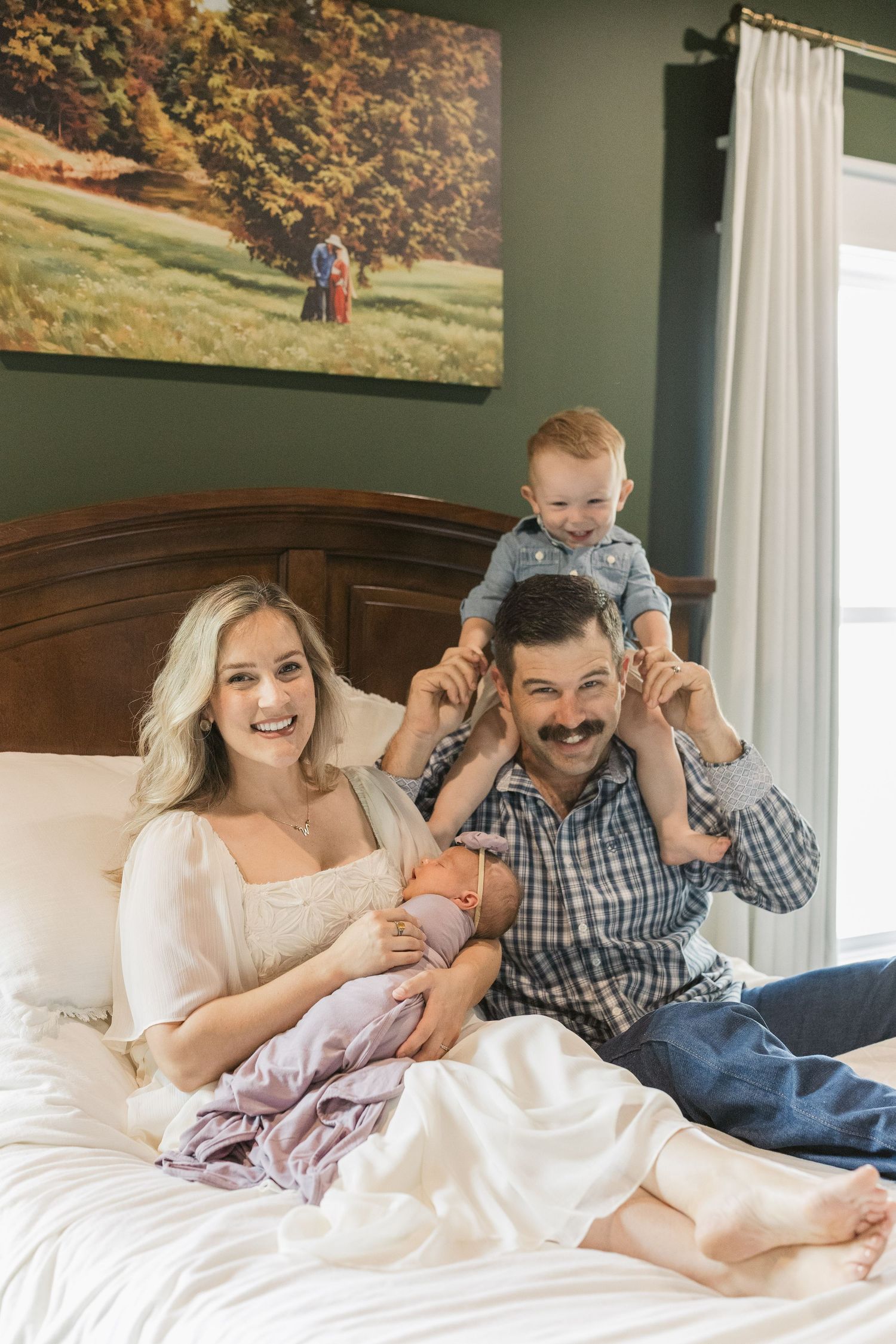 Smiling family portrait on bed, mother holding newborn as father balances toddler on shoulders.