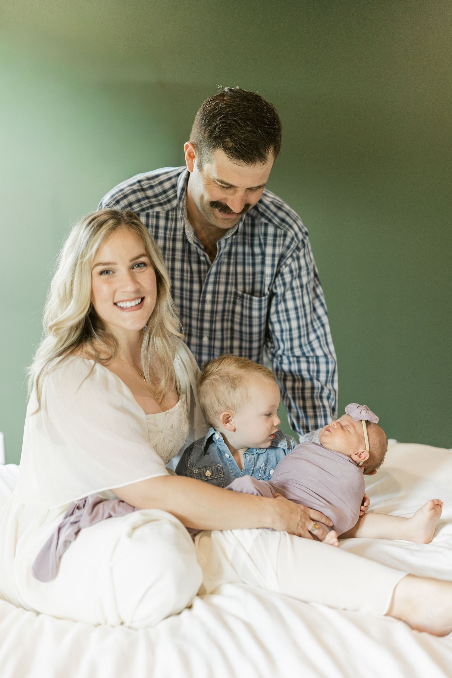 Happy family with parents, toddler, and newborn baby posing together on a white bed against a green wall.