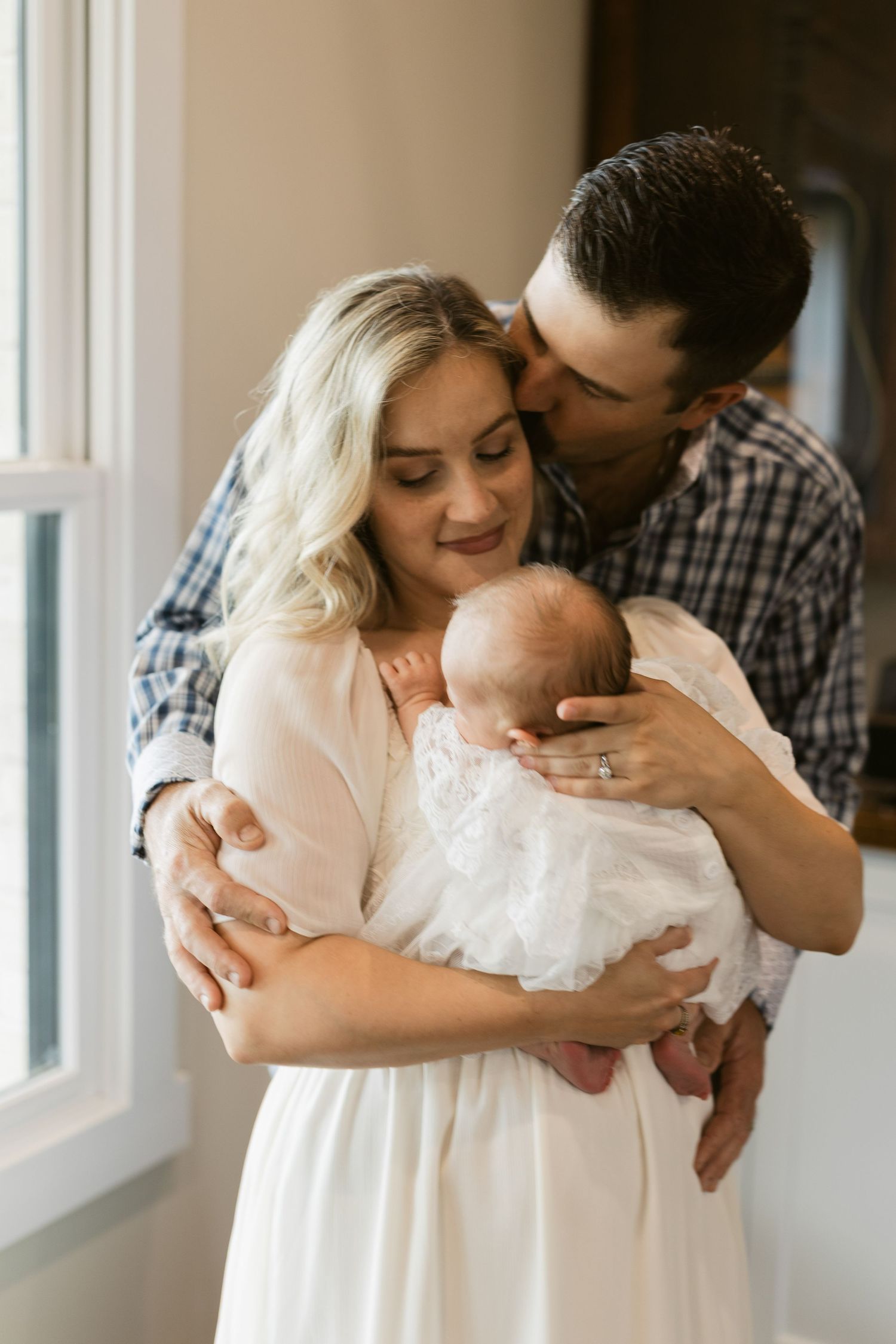 New parents lovingly hold their newborn baby, father kissing mother's head.