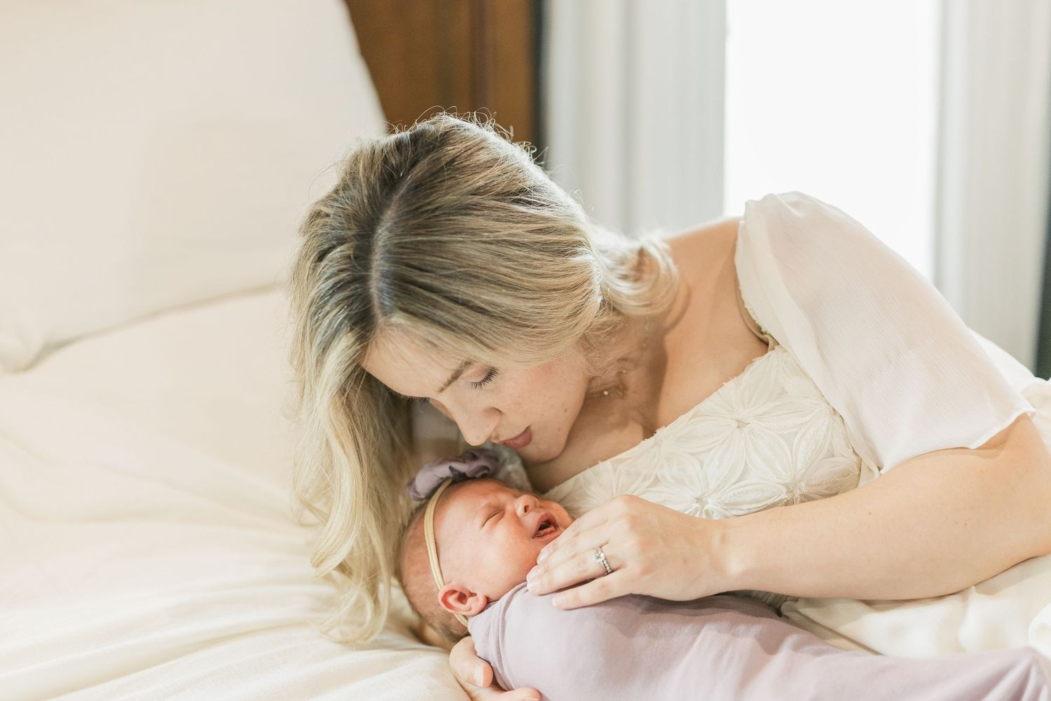 A tender mother gazes lovingly at her newborn baby wrapped in pink on a white bed.