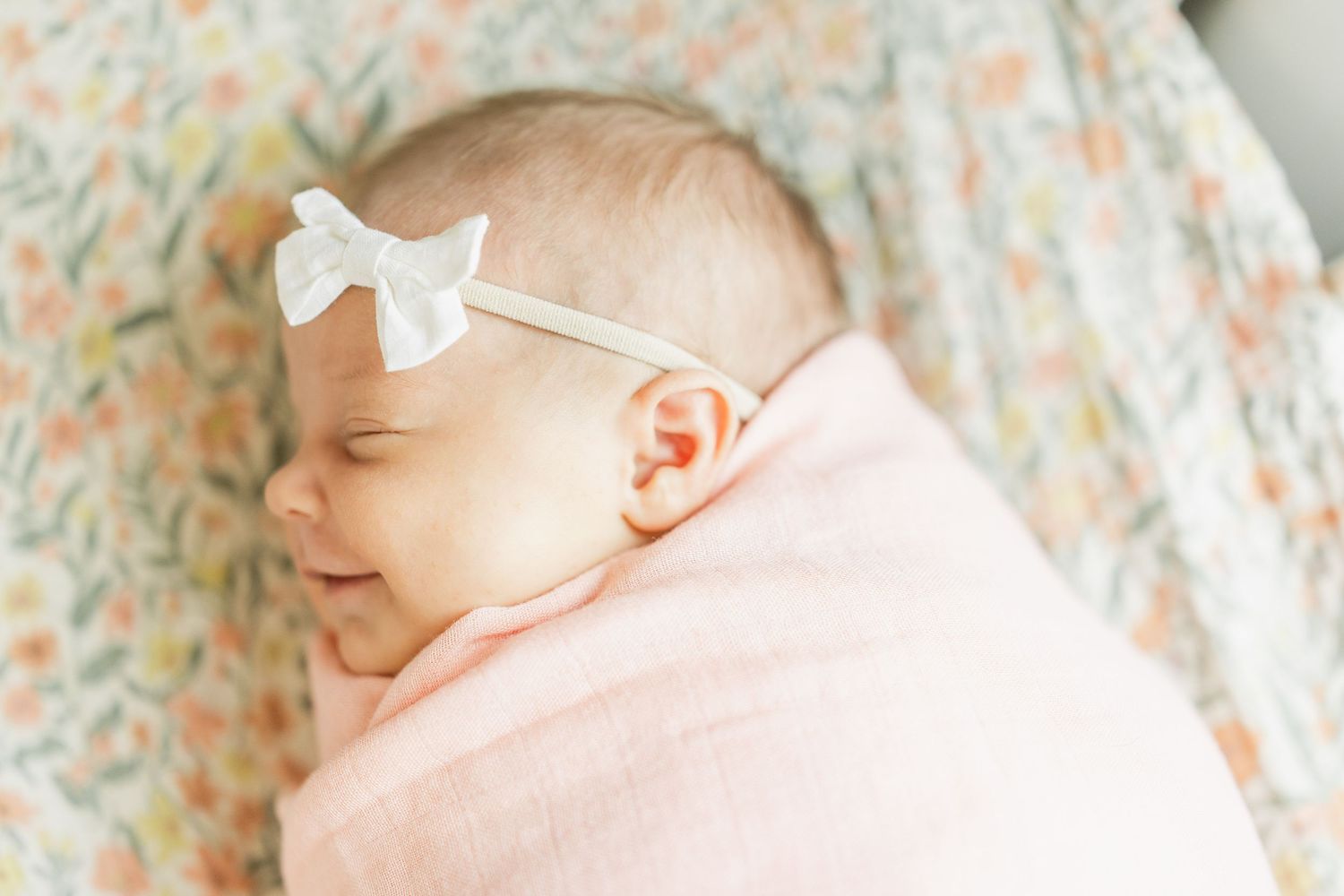 Sleeping newborn baby girl swaddled in pink blanket wearing a white bow headband.