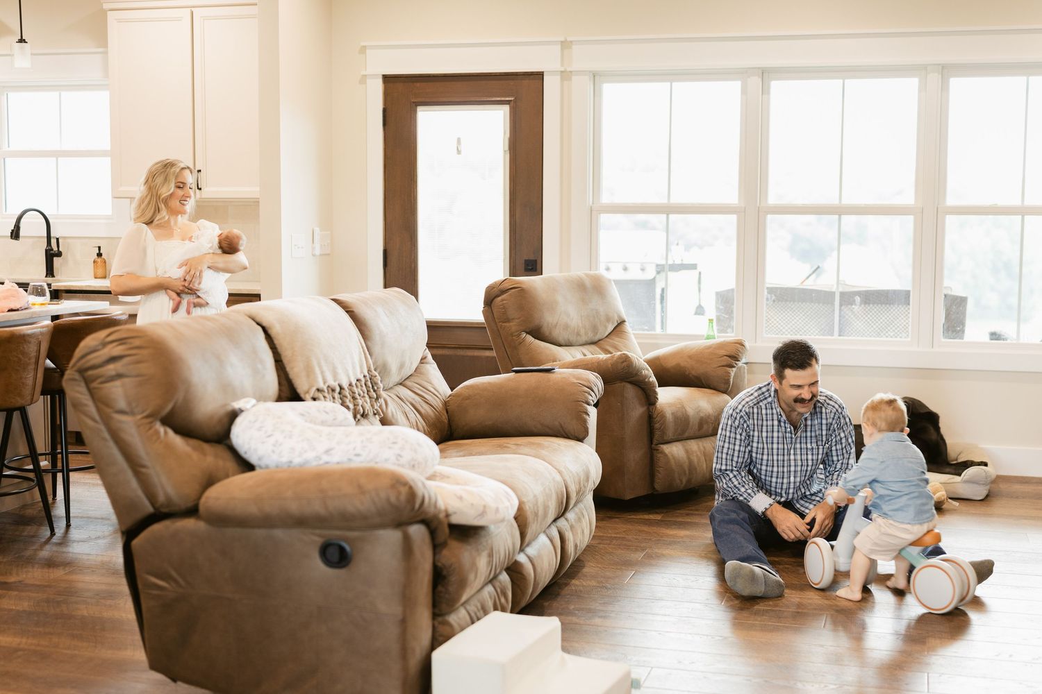 Family relaxing in a cozy living room with brown leather sofas, father playing with child on hardwood floor.