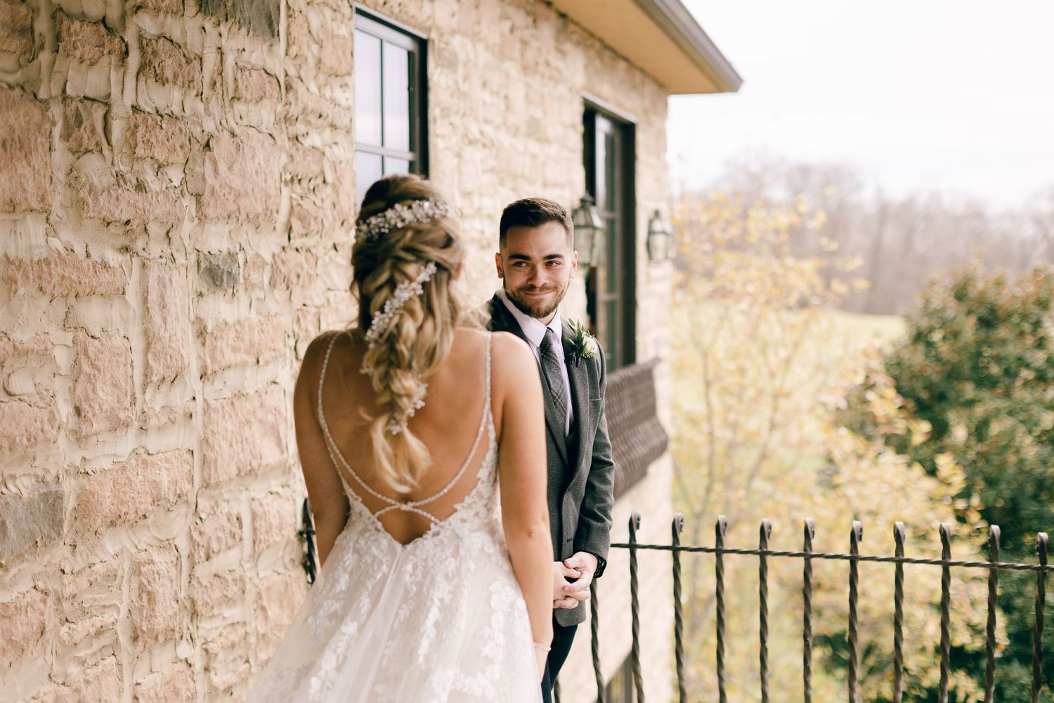 Bride and groom sharing a romantic moment on a stone building balcony, bride in backless lace gown with braided updo.