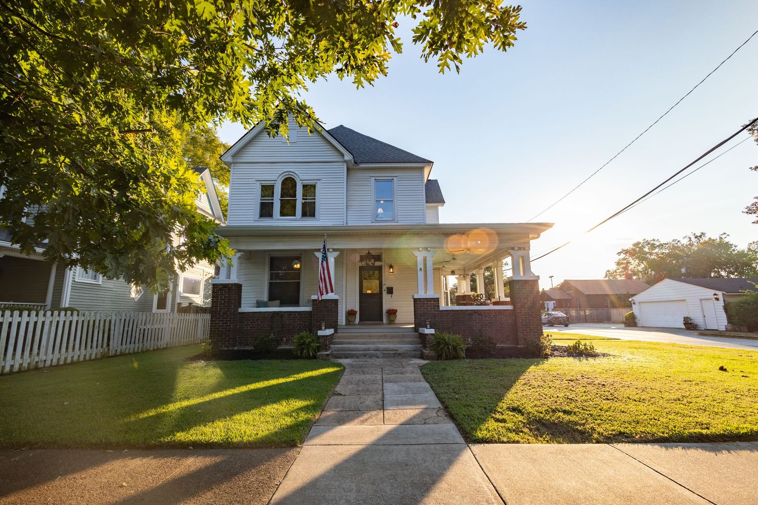 Classic two-story American home with brick foundation, covered porch, manicured lawn, and mature shade trees.