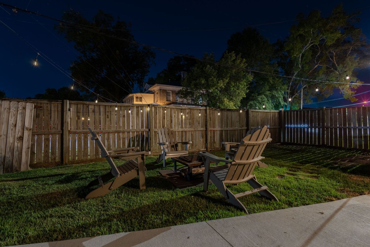 Cozy backyard fire pit area with Adirondack chairs, string lights, and wooden privacy fence at night.