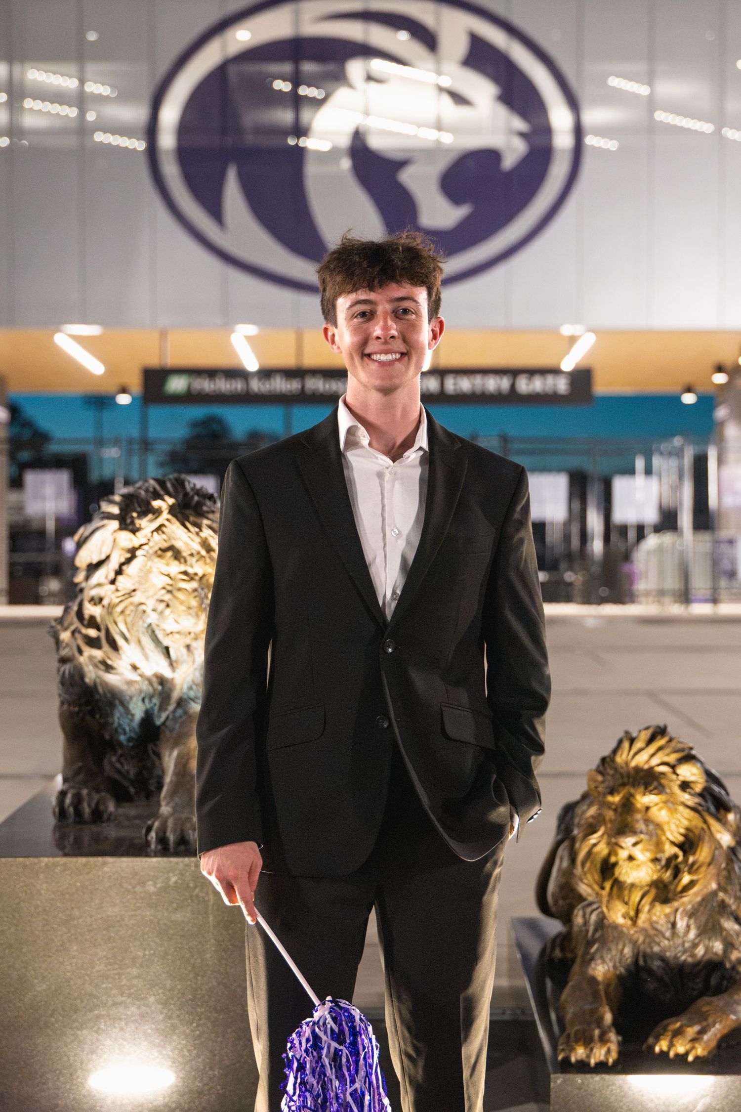 Young man in black suit smiling beside golden lion statues at a university arena entry gate.