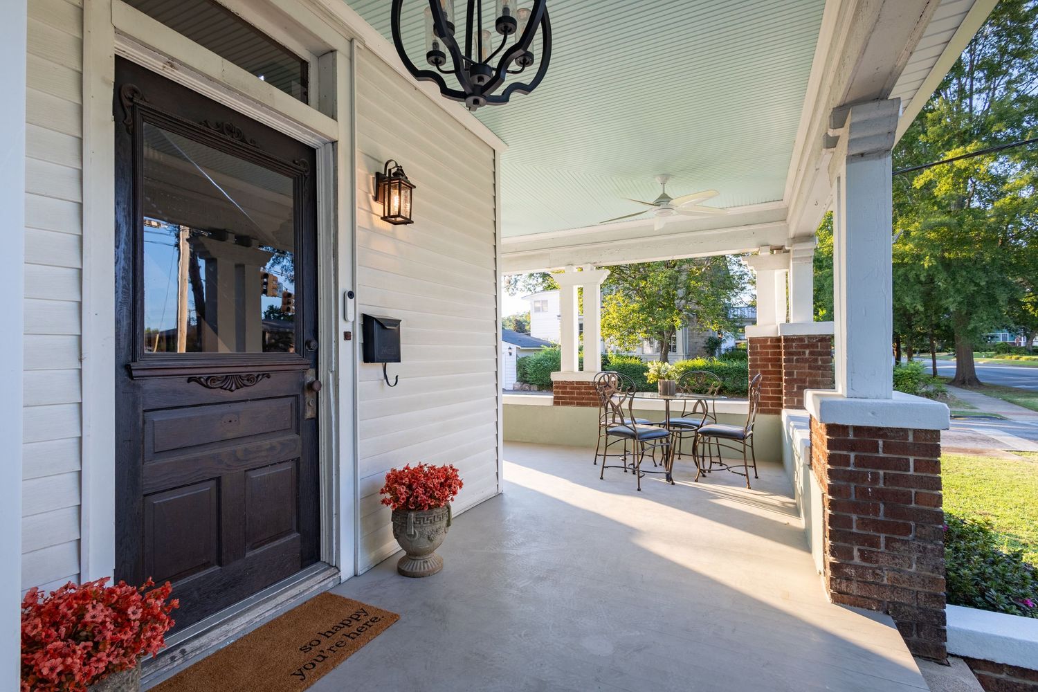 Charming home front porch with dark door, brick columns, chandelier lighting, and red potted flowers.