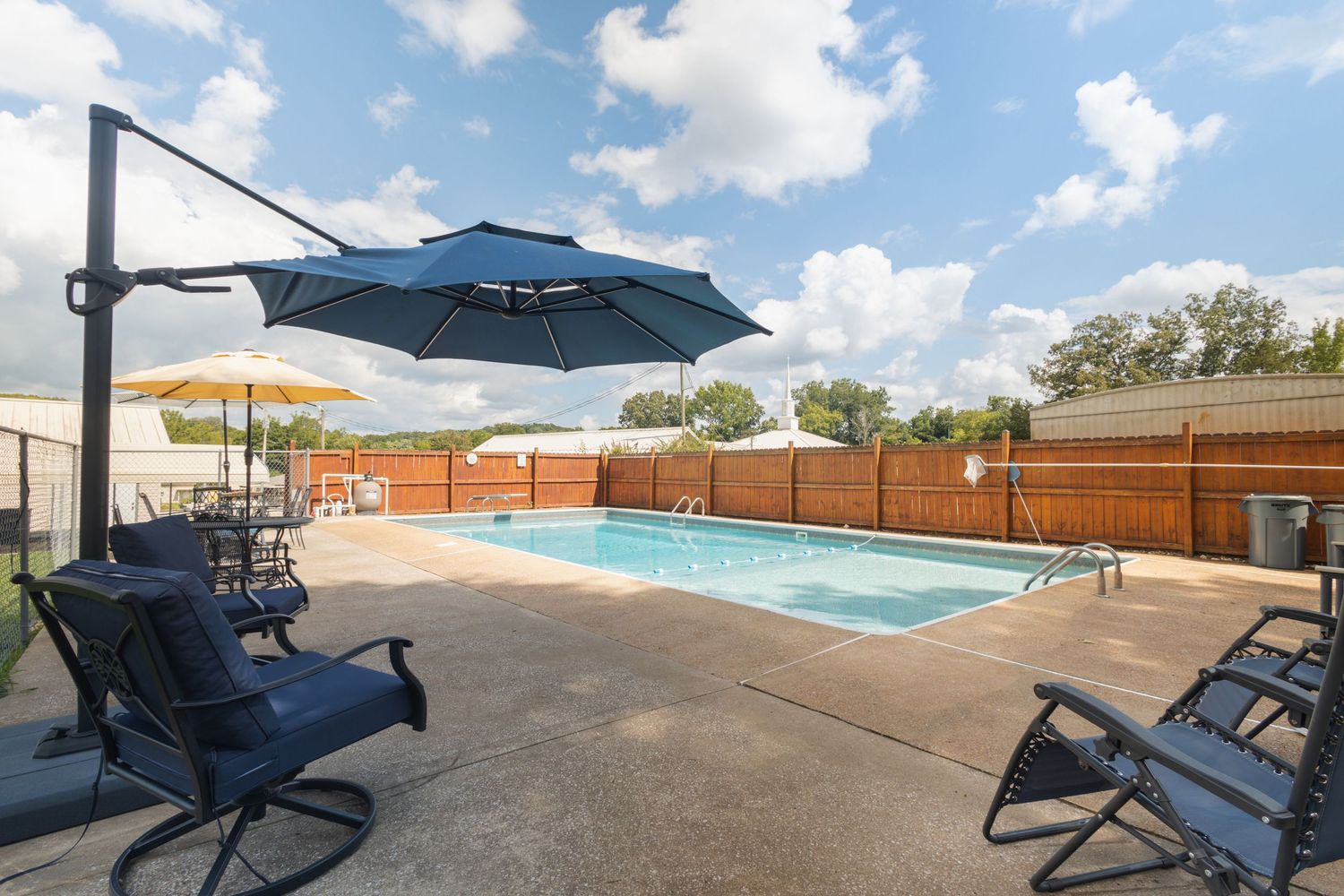 Outdoor pool area with blue lounge chairs, cantilever umbrellas, and wooden privacy fencing.