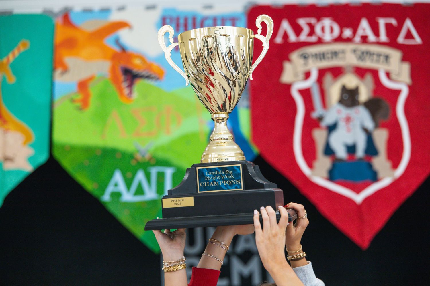Hands raising a gold trophy cup in front of colorful Greek organization banners at campus event.
