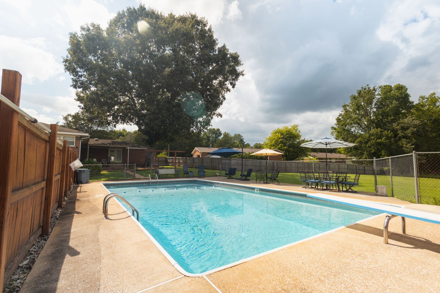 Backyard swimming pool with clear blue water surrounded by green lawn under dramatic cloudy sky.