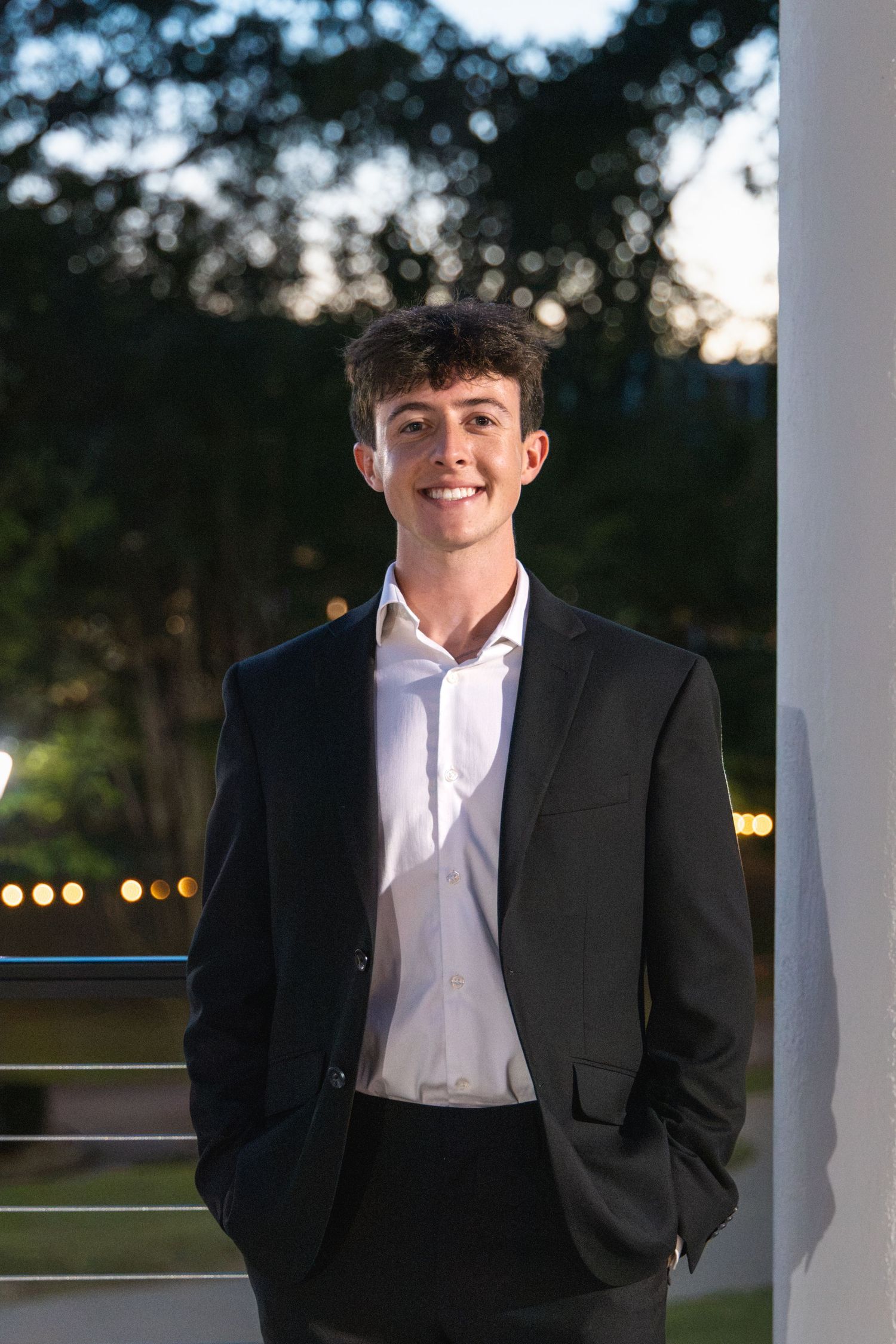 Smiling young man in black suit and white shirt posing outdoors at dusk near a white column.