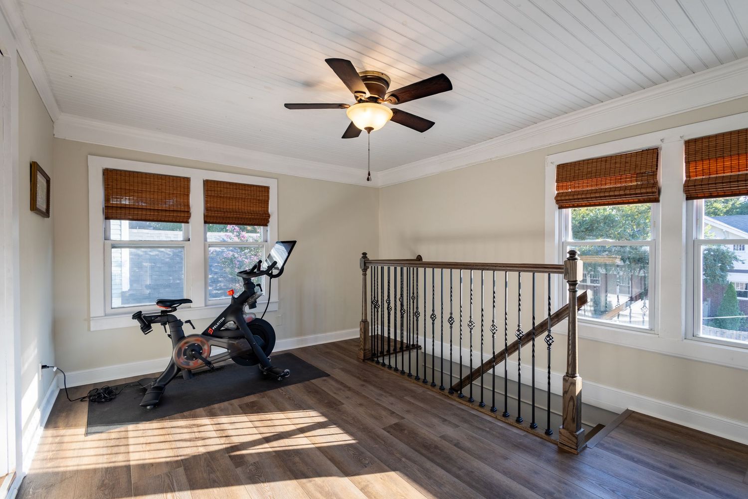Bright upper landing with exercise bike, ceiling fan, hardwood floors, and wooden staircase railing.