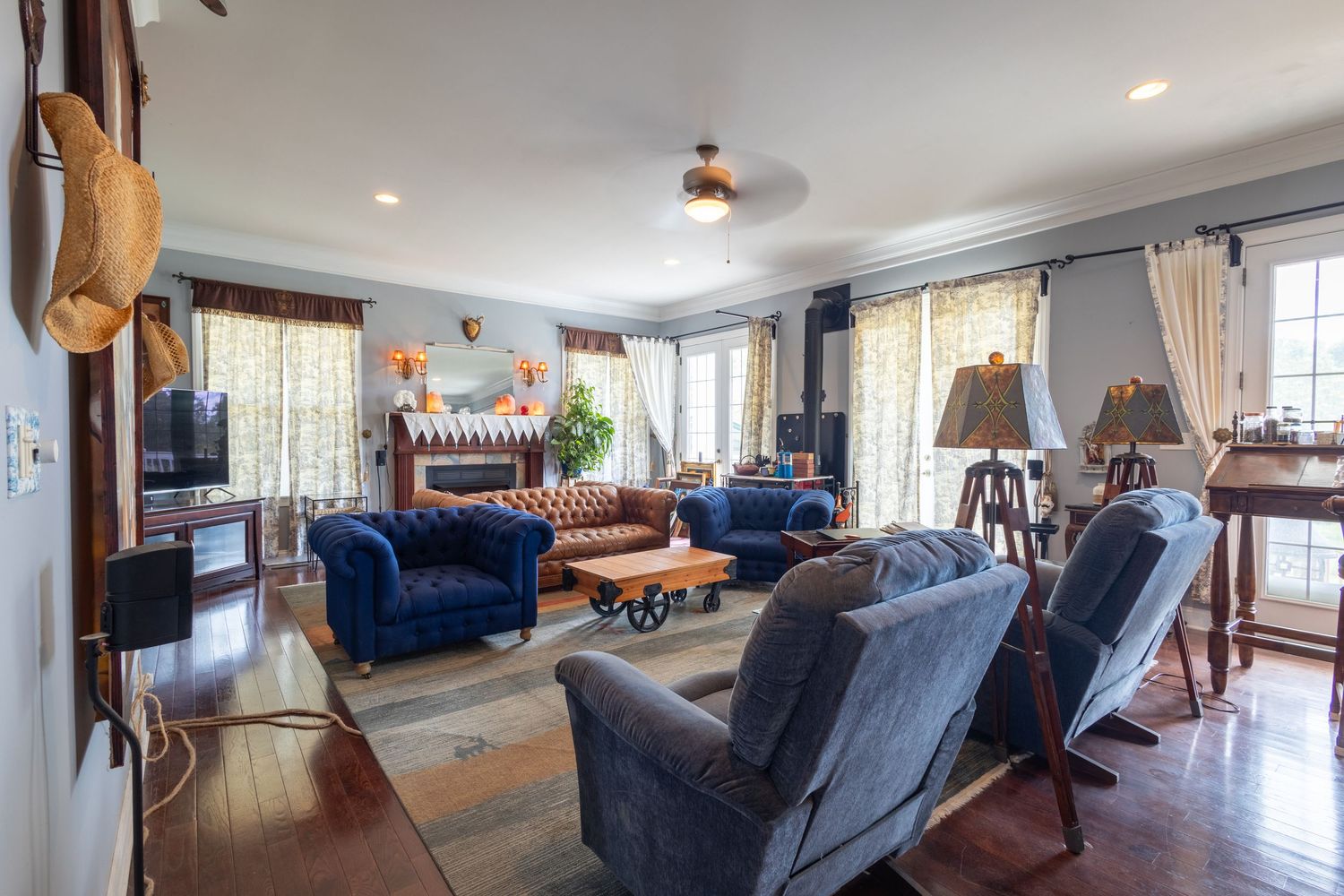 Spacious living room with blue velvet sofas, hardwood floors, ceiling fan, and large windows letting in natural light.
