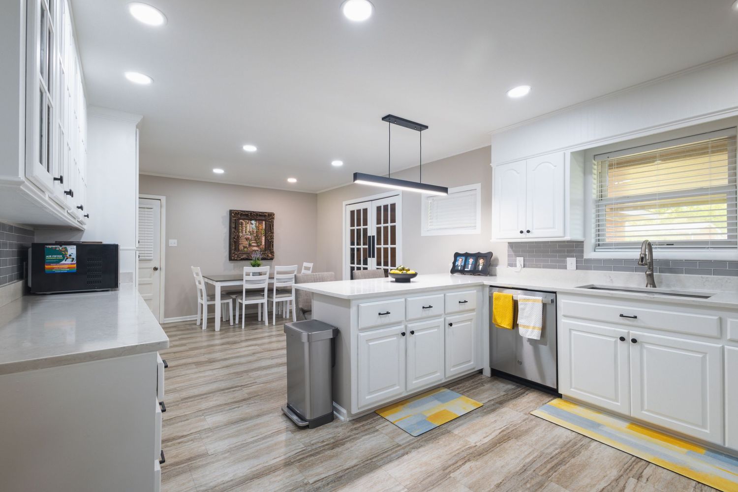Modern white kitchen with island, recessed lighting, hardwood floors, and open dining area beyond.