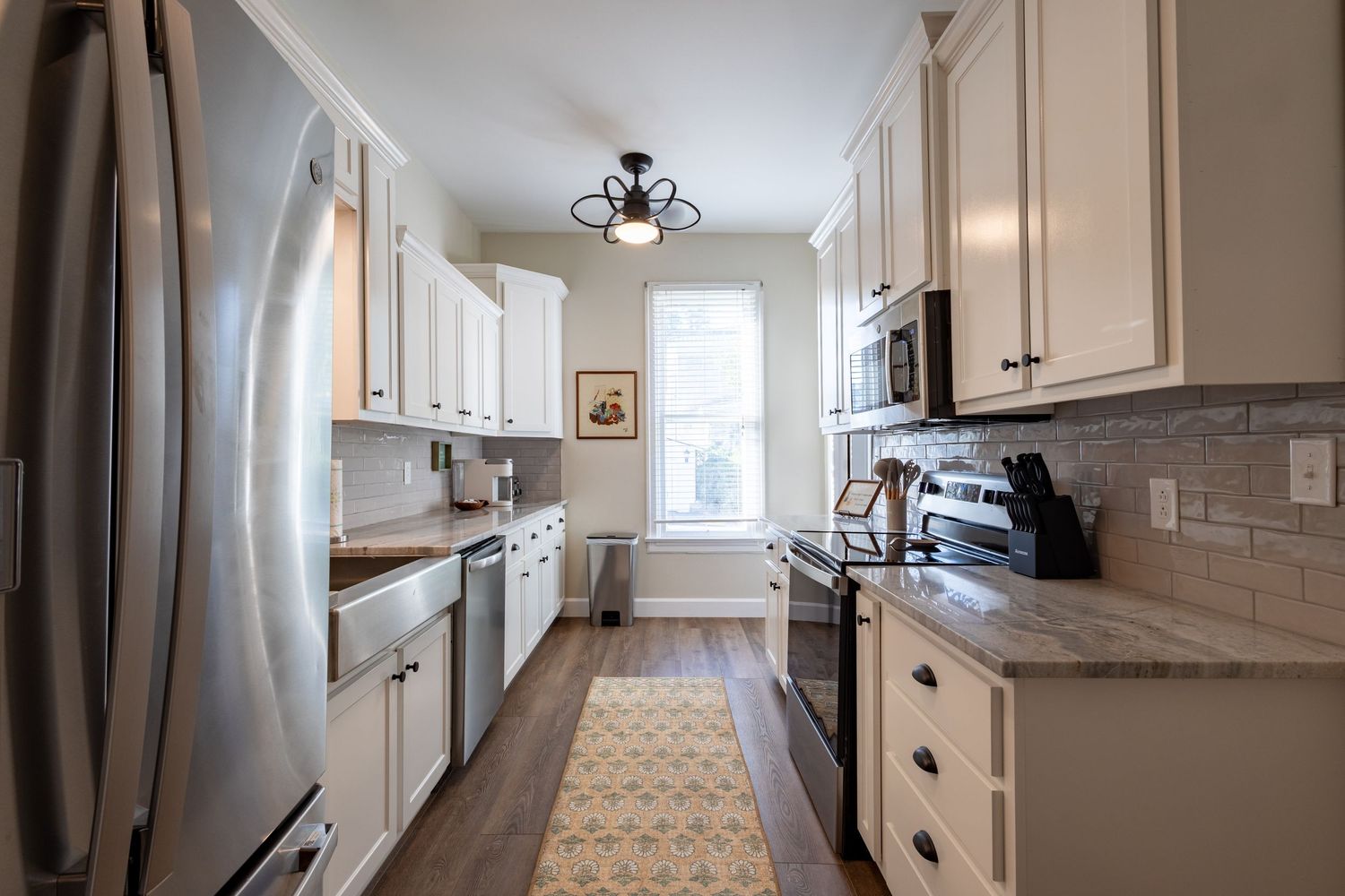 Narrow galley kitchen featuring white cabinetry, granite countertops, stainless appliances, and a patterned runner rug.