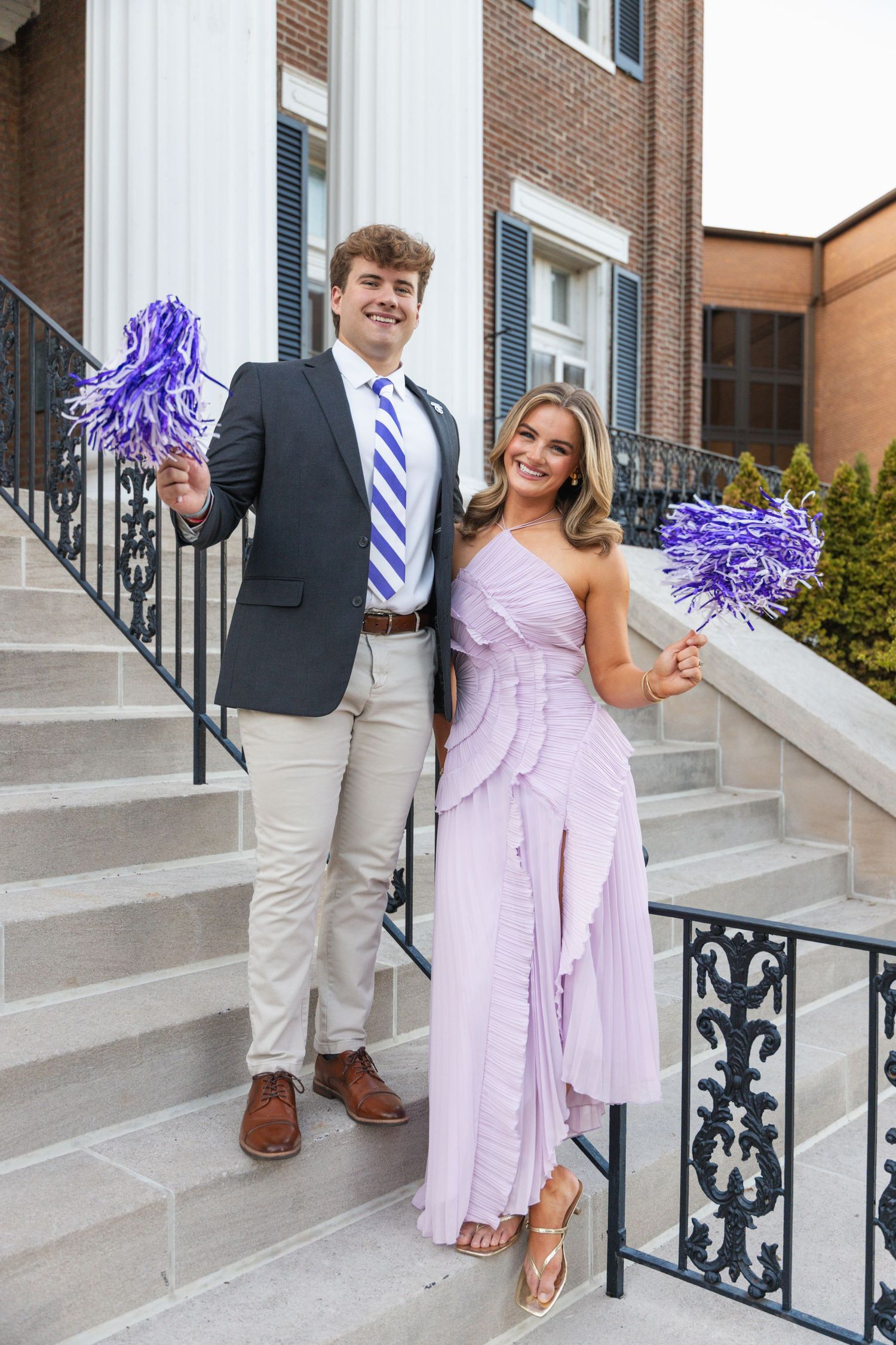 Couple dressed formally holding purple pom-poms on brick building steps at college homecoming event.