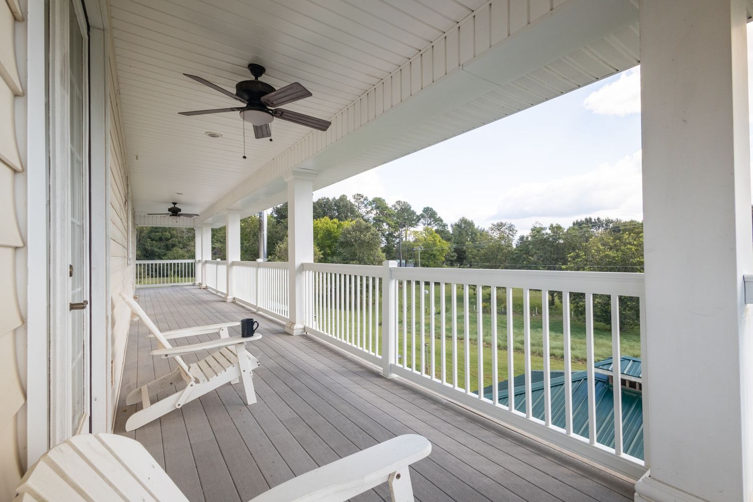 Covered porch with white Adirondack chairs, ceiling fan, white railings, and scenic green yard view.