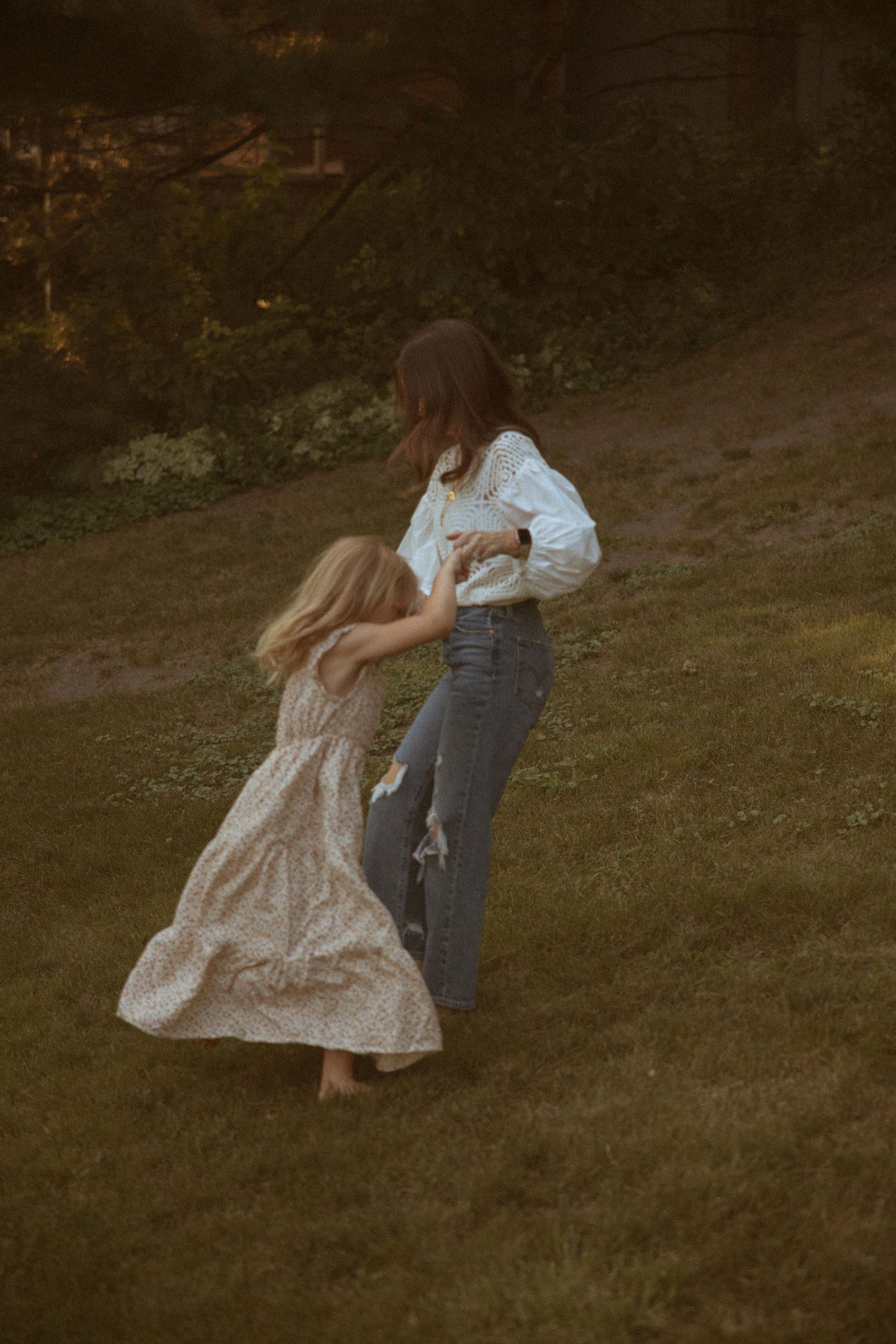 A girl in a floral dress twirls barefoot on a grassy lawn while a woman in jeans and a white blouse holds her hands at golden hour.