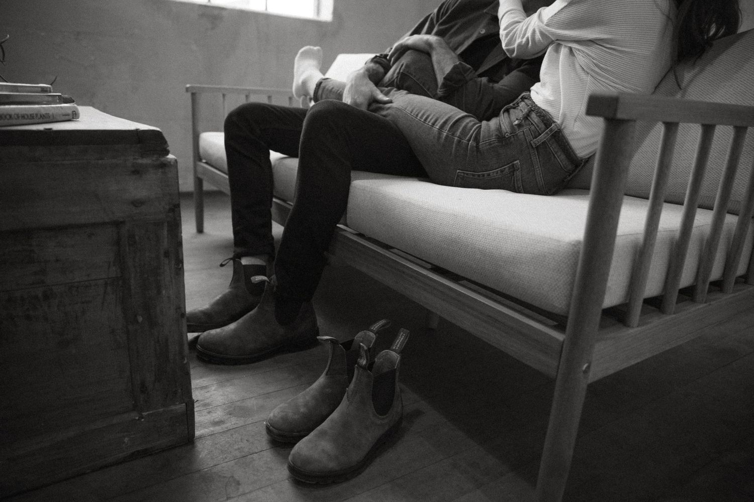 Black and white photo of a person seated on a bench with worn leather boots on the wooden floor beside a rustic trunk.