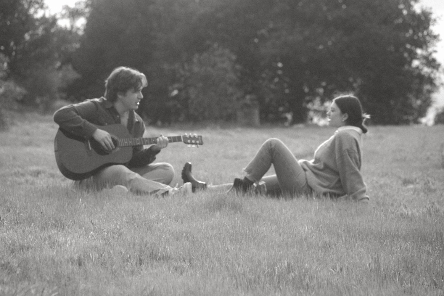 Black and white photo of a young man playing acoustic guitar while a woman relaxes on the grass in a park.