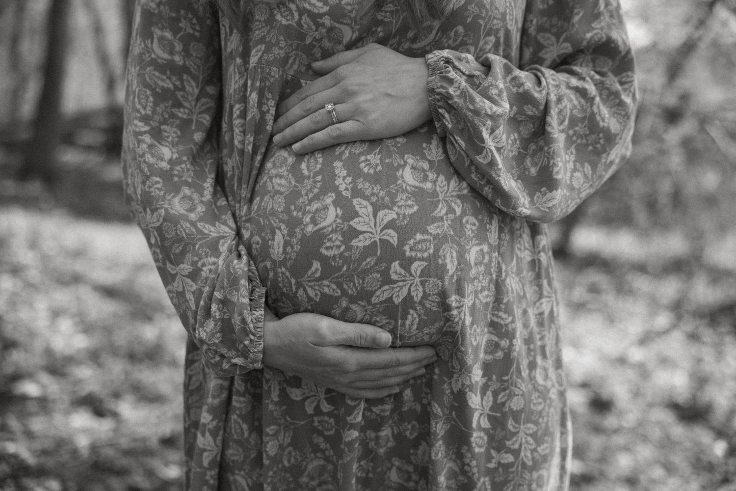 Black and white photo of a pregnant woman in a floral dress cradling her baby bump in a sunlit woodland.