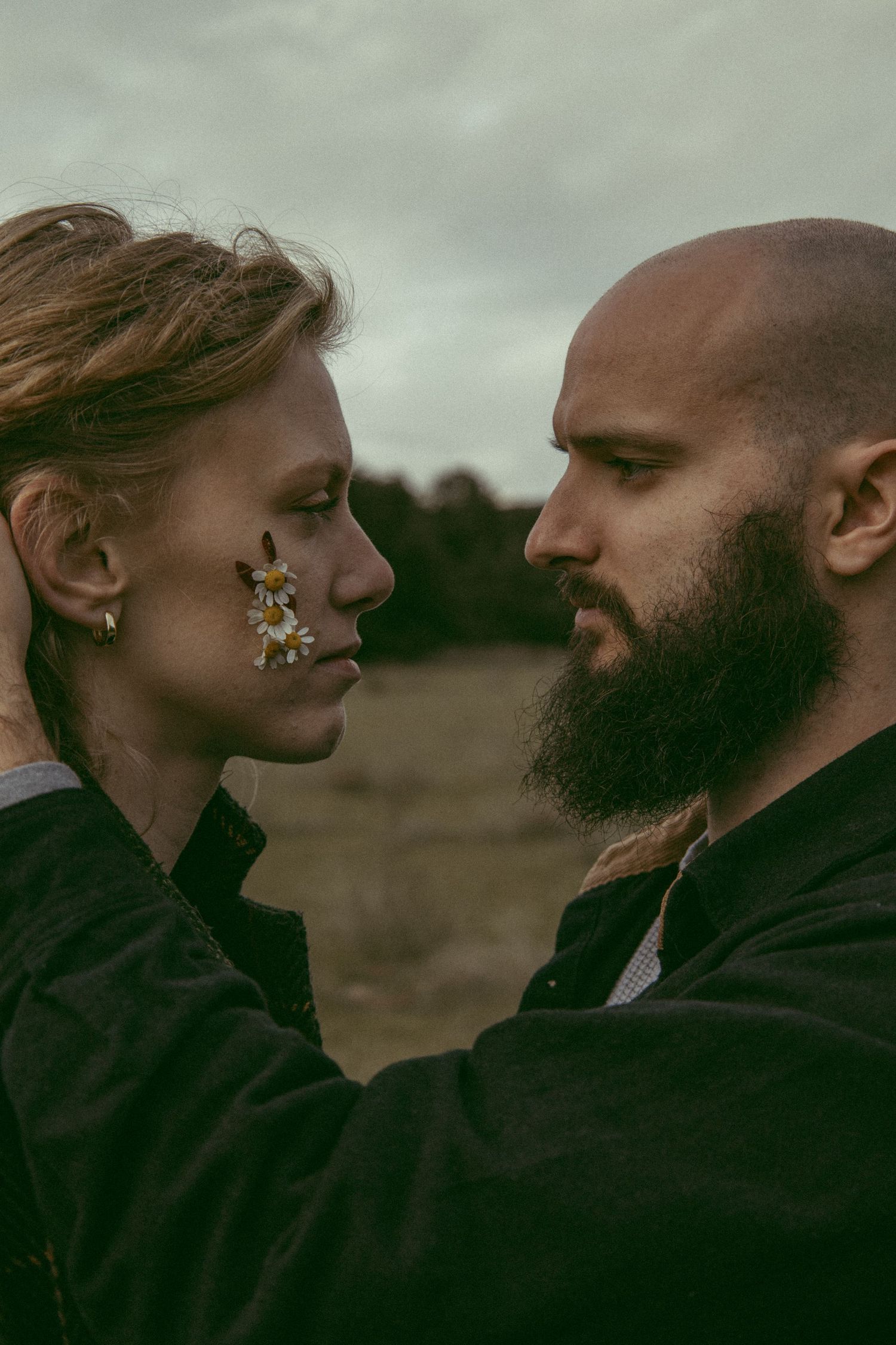 Moody portrait of a bearded man and a woman with small daisies on her cheek standing face to face in an open field.