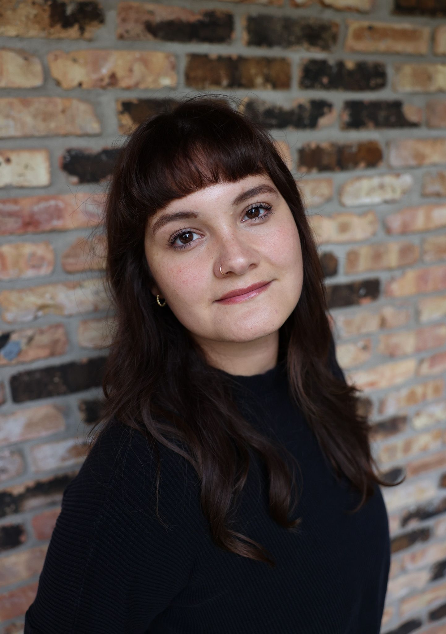 A young woman with long dark hair and bangs smiles softly while posing in front of a rustic exposed brick wall.